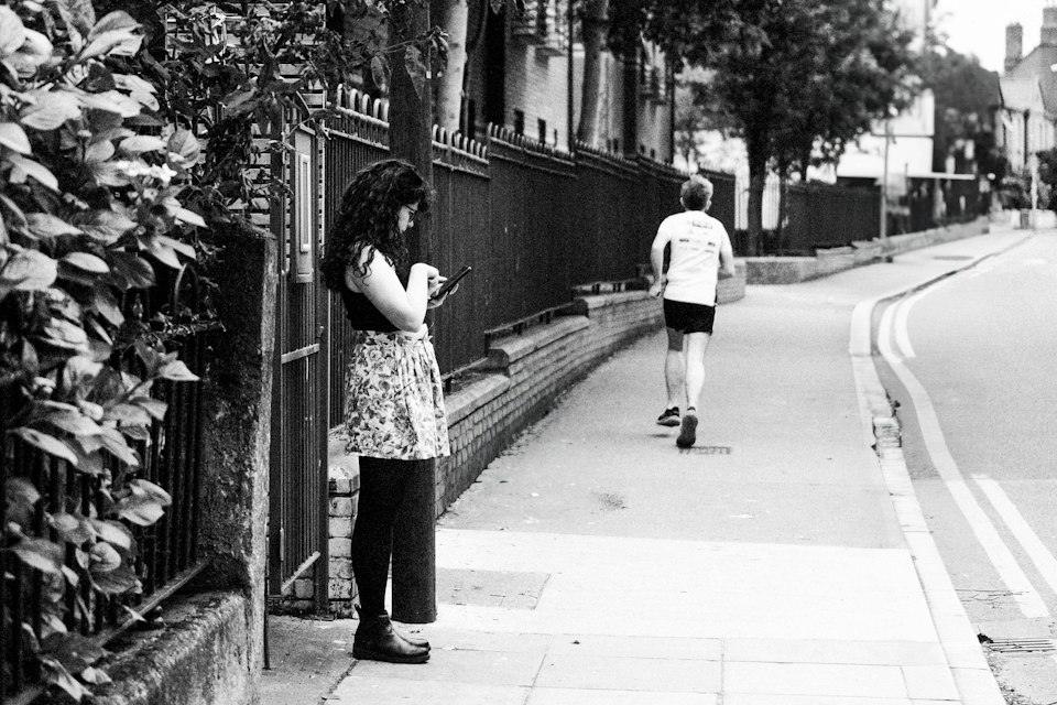 Woman on sidewalk looking at phone while man jogs past
